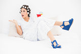 Brunette in hair rollers and wedge shoes holding a cocktail gesturing on bed