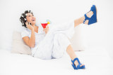 Brunette in hair rollers and wedge shoes holding a cocktail making a call on bed