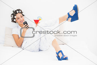Brunette in hair rollers and wedge shoes having a cocktail sending a text on bed