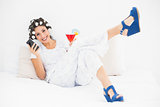 Brunette in hair rollers and wedge shoes having a cocktail using smartphone on bed