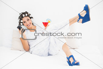 Brunette in hair rollers and wedge shoes having a cocktail using smartphone on bed