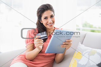 Smiling brunette sitting on her sofa using tablet to shop online