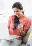 Cheerful brunette sitting on her sofa using laptop to shop online