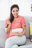 Cheerful brunette sitting on her sofa eating bowl of popcorn