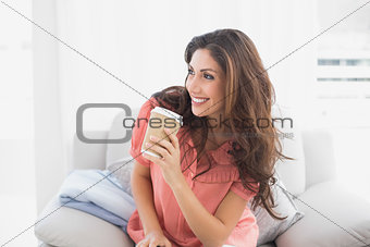 Happy brunette sitting on her sofa holding disposable cup