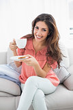 Smiling brunette sitting on her sofa holding cup and saucer