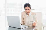 Serious businesswoman reading newspaper while working on laptop