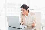 Stern businesswoman holding newspaper while working on laptop