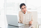 Stern businesswoman holding newspaper while working on laptop looking at camera
