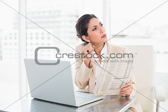 Stern businesswoman holding newspaper while working on laptop looking away