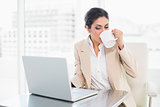 Cheerful businesswoman drinking from mug while working on laptop
