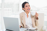 Laughing businesswoman holding cup while working on laptop