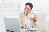 Happy businesswoman holding cup while typing on laptop