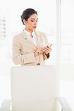 Frowning businesswoman standing behind her chair texting on her phone