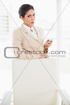 Frowning businesswoman standing behind her chair holding her phone glaring at camera