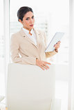 Frowning businesswoman standing behind her chair holding tablet pc