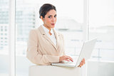 Smiling businesswoman standing behind her chair holding laptop