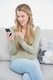 Smiling pretty blonde text messaging while sitting on cosy sofa