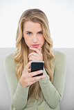 Thoughtful pretty blonde sending a text sitting on cosy sofa