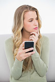 Pensive pretty blonde sending a text sitting on cosy sofa
