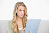 Focused pretty blonde using her tablet sitting on cosy sofa