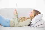 Happy pretty blonde using her tablet lying on cosy sofa