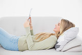 Smiling pretty blonde using her tablet lying on cosy sofa