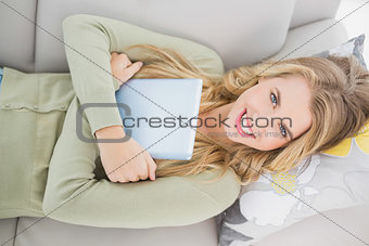 Overhead of pretty blonde holding her tablet lying on cosy sofa
