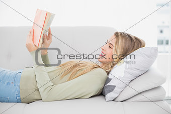 Smiling pretty blonde reading book lying on cosy sofa