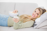 Cheerful pretty blonde holding popcorn lying on cosy sofa