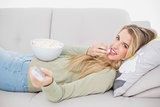Cheerful pretty blonde eating popcorn lying on cosy sofa