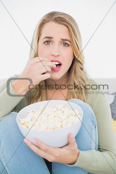 Surprised pretty blonde eating popcorn sitting on cosy sofa