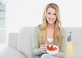 Smiling pretty blonde eating strawberries sitting on cosy sofa
