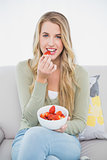 Cheerful pretty blonde eating strawberries sitting on cosy sofa