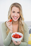 Happy pretty blonde eating strawberries sitting on cosy sofa