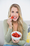 Happy cute blonde eating strawberries sitting on cosy sofa