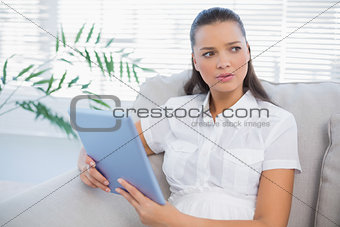 Thoughtful cute woman using tablet sitting on cosy sofa