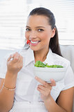 Cheerful pretty woman eating healthy salad sitting on sofa