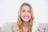 Happy cute blonde eating cookie with milk sitting on cosy sofa