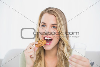 Cheerful cute blonde eating cookie with milk sitting on cosy sofa