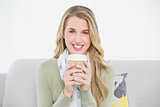 Cheerful cute blonde holding coffee sitting on cosy sofa