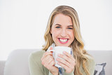 Smiling cute blonde holding cup of coffee sitting on cosy sofa