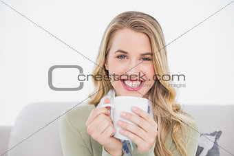 Smiling cute blonde holding cup of coffee sitting on cosy sofa