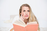 Thoughtful gorgeous model lying on cosy bed holding book
