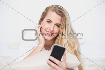 Smiling model holding her smartphone lying on cosy bed