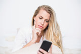 Frowning model holding her smartphone lying on cosy bed