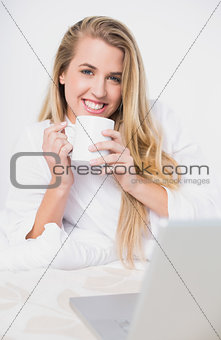 Smiling pretty model holding coffee lying on cosy bed