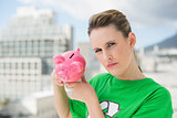 Serious woman wearing green recycling tshirt holding piggy bank