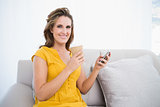 Smiling woman sitting on sofa holing coffee and using phone