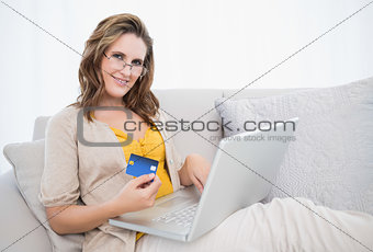 Smiling woman lying on sofa showing credit card holding laptop